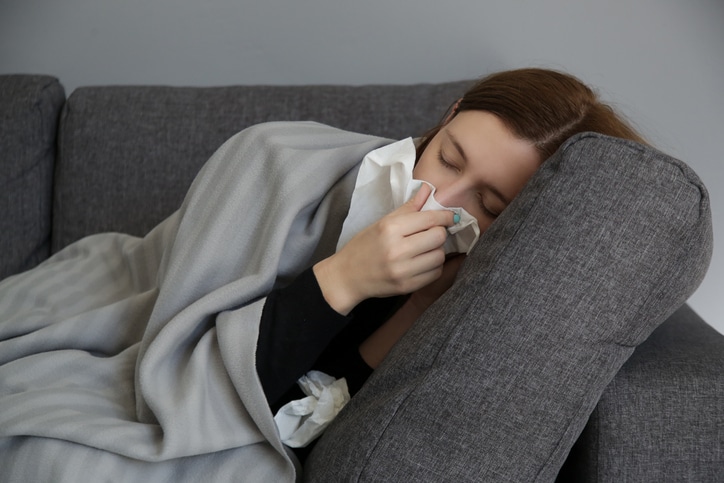 Young woman on the sofa blowing nose into a white paper tissue. Studio shot of young woman with allergy symptoms sneezing into a tissue. Flu, cold or allergy symptom.