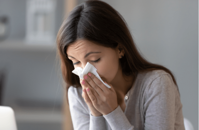 Sick young woman sitting indoors holding tissue handkerchief blowing running nose
