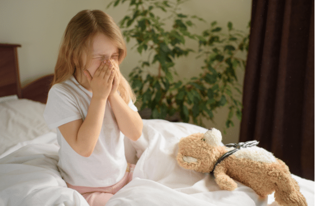 A little girl sits and sneezes on the bed among the blankets and pillows.